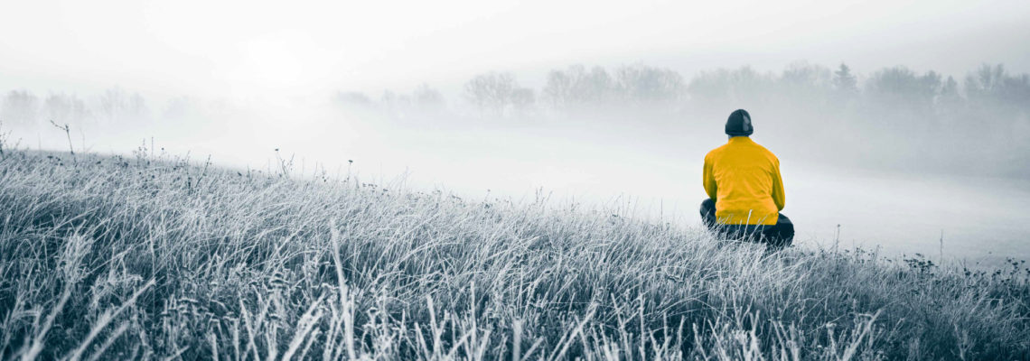 Foto av en åker i frost. Til høyre for bildet, omtrent i midten dybdemessig, sitter enmann på huk og ser videre inn i bildet. Han ser på en tåkebelagt åker og en skoglinje i det fjerne. Fotoet er i blågrå toner. Unntaket er jakken til mannen, som er i en sterk guloransje farge.