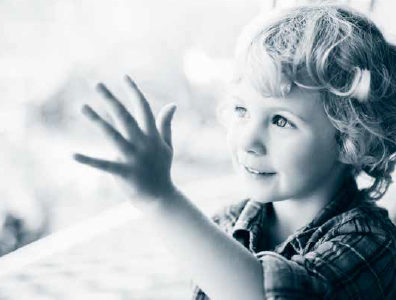 Foto i gråblå toner av en liten gutt med kort, krøllete hår som står ved et vindu og er ut av det mens han har hånden på glasset.