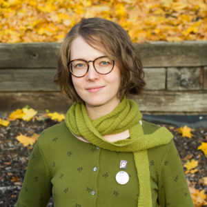 Fotograif av kvinne med grønn cardigan, grønt, tynt skjerf, brunt år til kjeven og runde briller som smiler og ser inn i kamera. I bakgrunnen er det oransje høstløv.