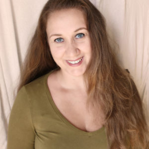 Foto av kvinne i grønn genser, langt, brunt hår og blå øyne som smiler og ser inn i kameraet. Bakgrunnen er et beige kanvas.