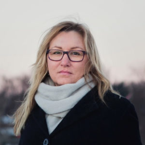 Fotografi av Marianne Kristiansdatter Finstad. En kvinne med langt, blond hår, briller, hvitt skjerf og sort kåpe ser inn i kamera med et nøyralt uttrykk.