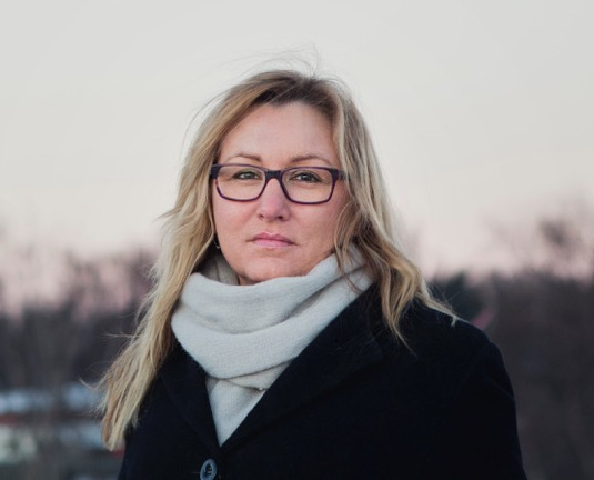 Fotografi av Marianne Kristiansdatter Finstad. En kvinne med langt, blond hår, briller, hvitt skjerf og sort kåpe ser inn i kamera med et nøyralt uttrykk.