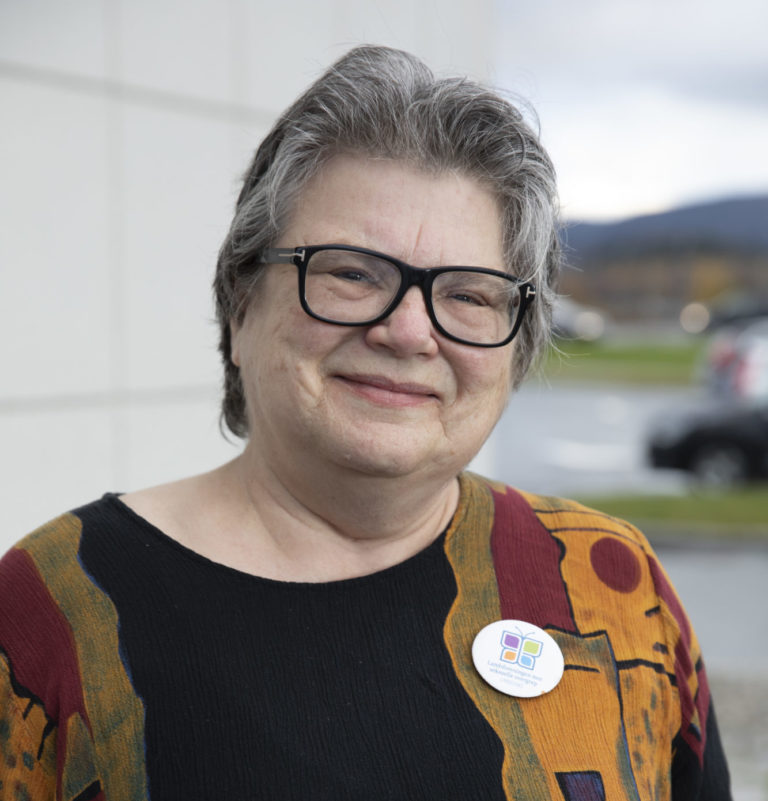 IngridJohanneVaalund Foto av kvinne med kort hår med gråtoner. Hun har på seg briller, smiler og ser rett i kamera. Hun har på seg en fargerik vest over en sort genser. Bakgrunne ner en blanding av et uklart landskap og en hvit vegg.