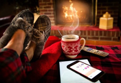 Fotografi. Fotoet viser to føtter med tøfler på som hviler på en rød pute. Ved siden av puta er det en kaffekopp med koppvarmer somer strikket i juleaktig mønster. Kaffekoppen står på et bord. På bordet ligger også en fjernkontroll, et nettbrett og en mobiltelefon.