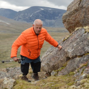 Foto av en mann i oransje boblejakke i et åpent fjellandskap. Mannen har proteser fra knærne og ned. Han klatrer i fjellet.