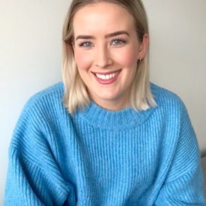 Ung kvinne med lyseblå, strikket genser og blondt hår til skuldrene smiler bredt og ser inn i kamera. Bak henne er en hvit vegg.