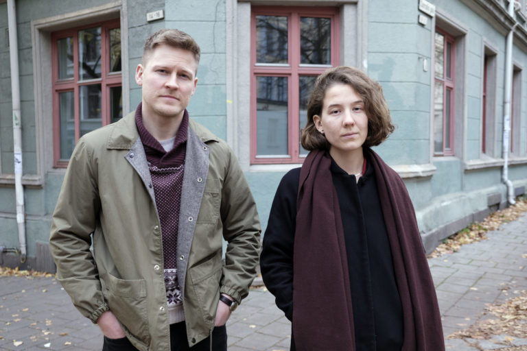 PressefotoRosenlundFilm En mann og en kvinne står foran et bygg og ser inn i kameraet. Mannen har grågrønn jakke og kortklipt hår. Kvinnen har sort jakke og lilla skjerf. De har en nøyral mine og ser rett i kamera. Bakgrunnen er et grønt bygg med vinduer med røde sprosser.