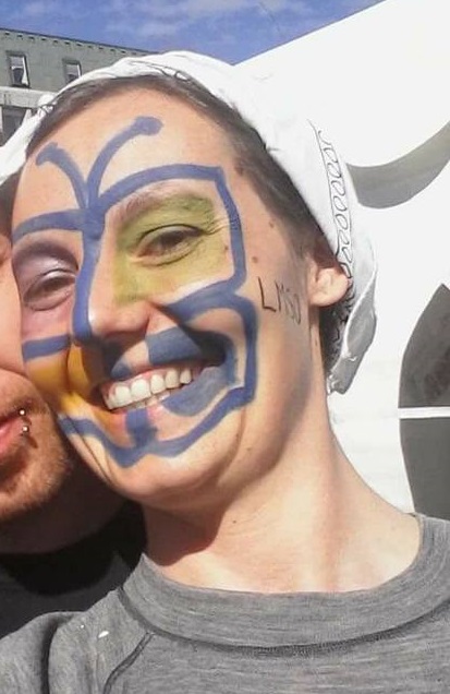 Foto av kvinne med ansiktsmaling. Ansiktsmalingen er logoen til LMSO: En somemrfugl i blått og med vinger i grønt, blått, lilla og oransje. Kvinenn smiler og er i kamera. På hodet har hun et hvitt skaut som dekker håret.