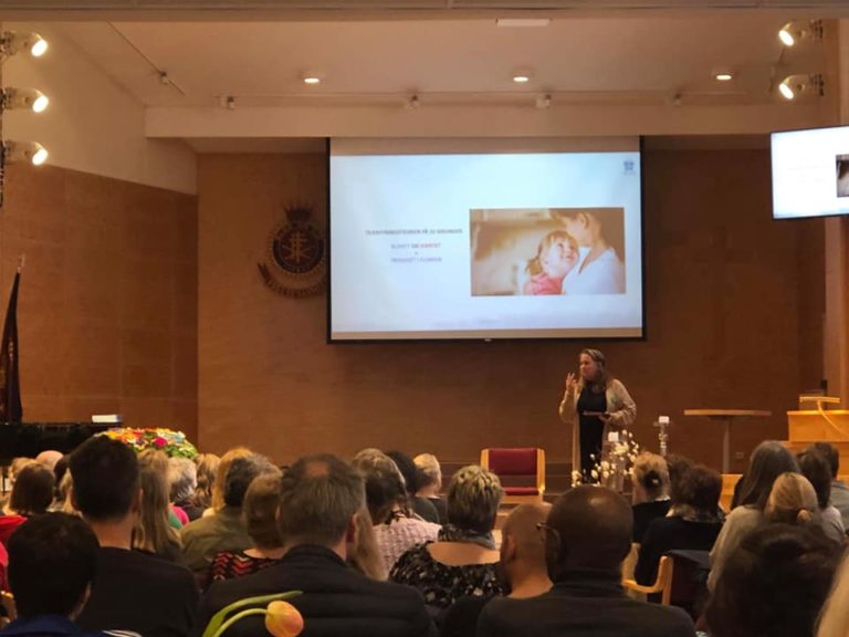 Verdighetsseminar Fotografi av forsamling, tatt bakfra. Forsamlingen ser på et lerret midt i rommet med tekst og bilde i en presentasjon, og en kvinne som står og prater foran lerretet.