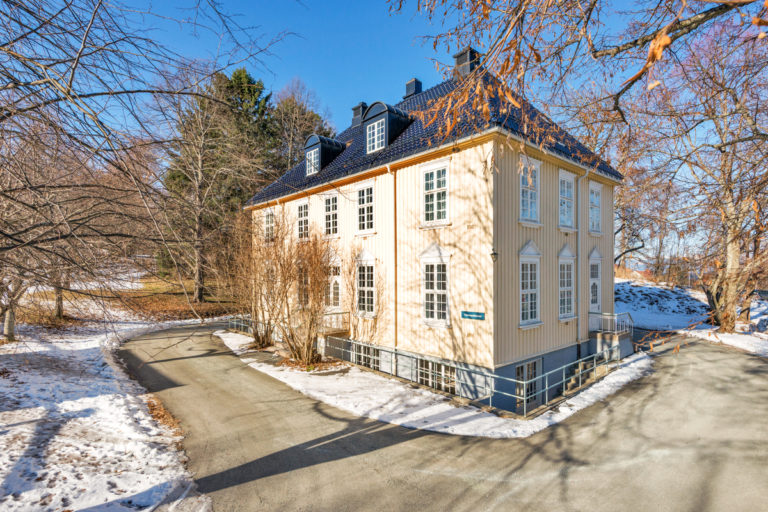 traumepoliklinikk Foto av et bygg malt i lys gul. Det er sprosser på vinduene, og ellers gammeldags stil. Det er blå himmel og sol og litt snø på omgivelsene.