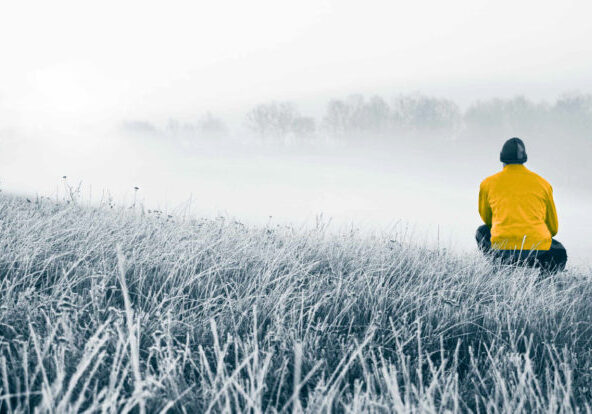 Foto av en åker i frost. Til høyre for bildet, omtrent i midten dybdemessig, sitter enmann på huk og ser videre inn i bildet. Han ser på en tåkebelagt åker og en skoglinje i det fjerne. Fotoet er i blågrå toner. Unntaket er jakken til mannen, som er i en sterk guloransje farge.