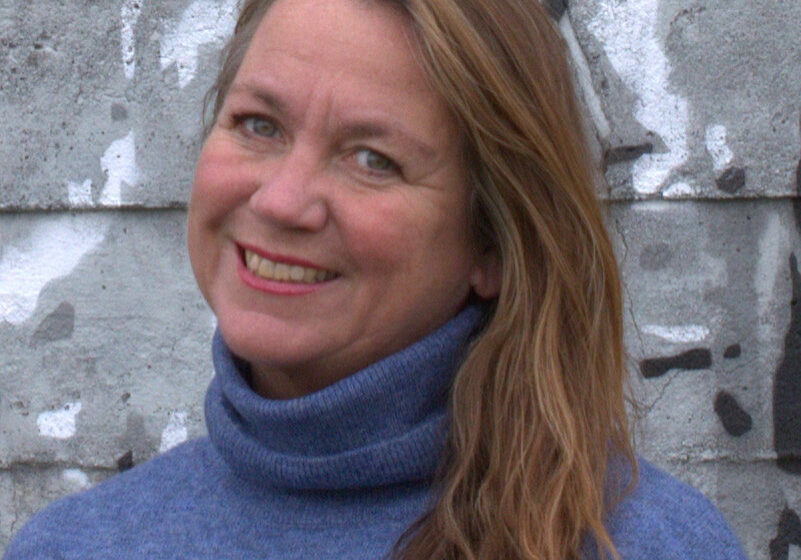 Foto av kvinne som smiler bredt og ser inn i kamera. Hun har lysebrunt/mellomblondt hår til skuldrene, blå øyne og en pologenser i en tåkeblå farge. Bakgrunnen er en røff betongvegg i grå med hvite og sorte malingsflekker på. Fotoet er tatt fra litt under skuldrene og opp.